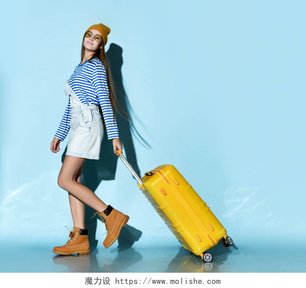 蓝色背景中一个时尚的女人拉着旅行箱全天候穿着牛仔裤、运动衫、太阳镜、靴子和帽子的少女。 笑着，背着蓝色背景的黄色行李箱走着.